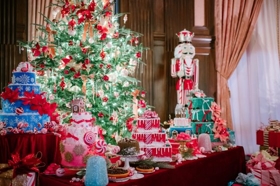  A table of faux-cakes, decorated for the holidays, in front of a decked out Christmas tree. 